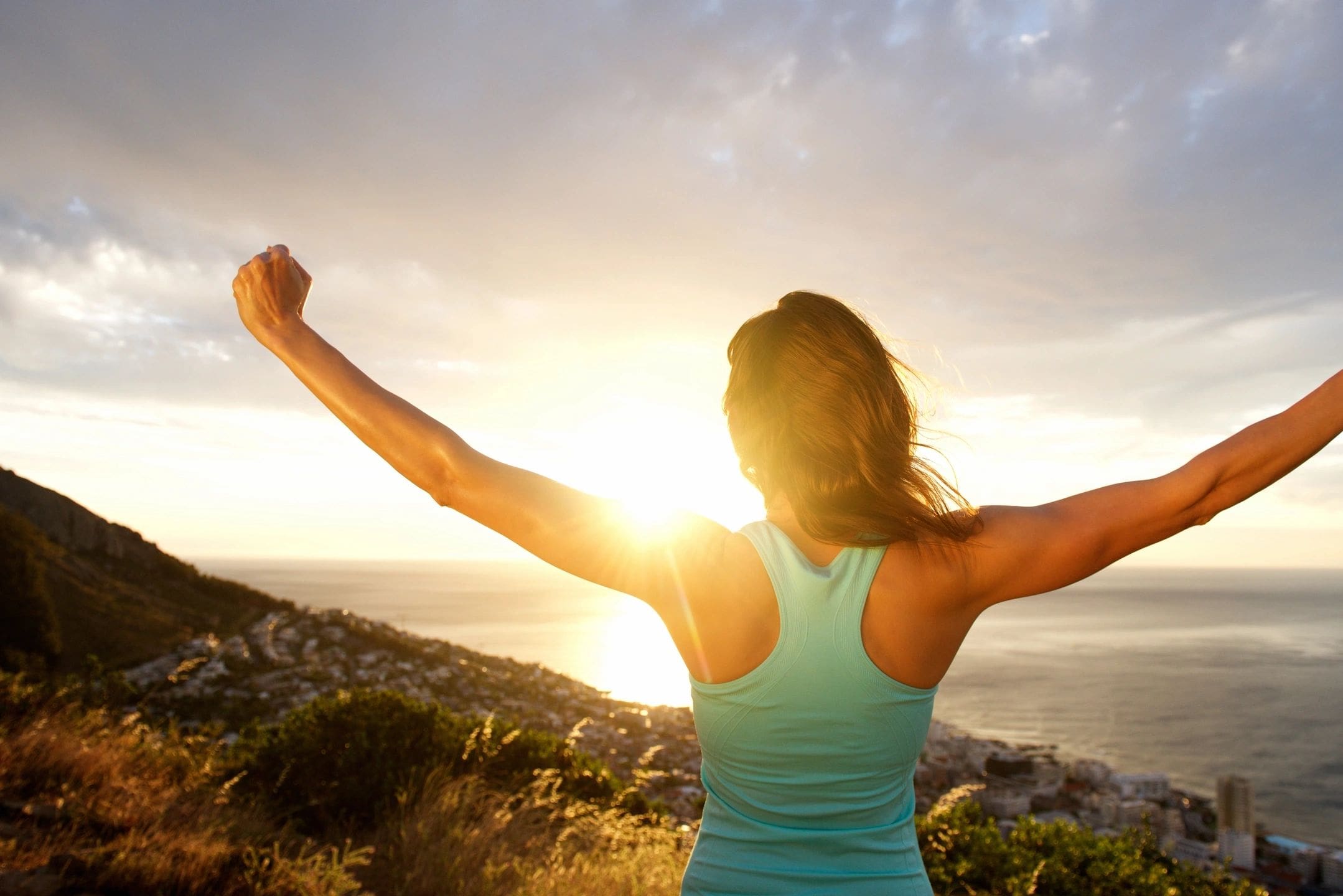 Woman in tank top with arms raised at sunset.